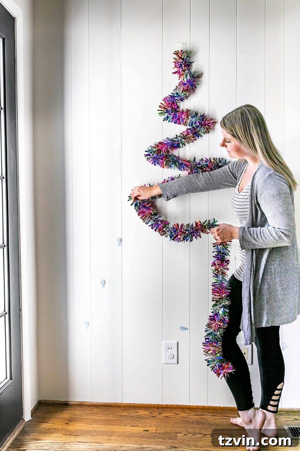 Process of hanging garland on strategically placed wall hooks to construct a DIY Christmas tree.