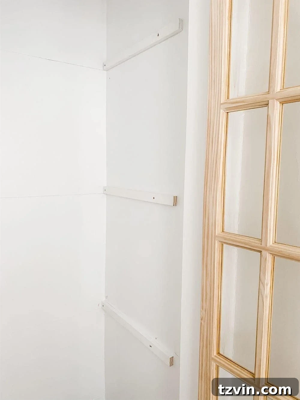 Simple Closet Transformation: Shelves and French Doors 11 A close-up of a person using a power drill to screw a 1x2 wooden board onto a closet wall, installing a shelf support brace.