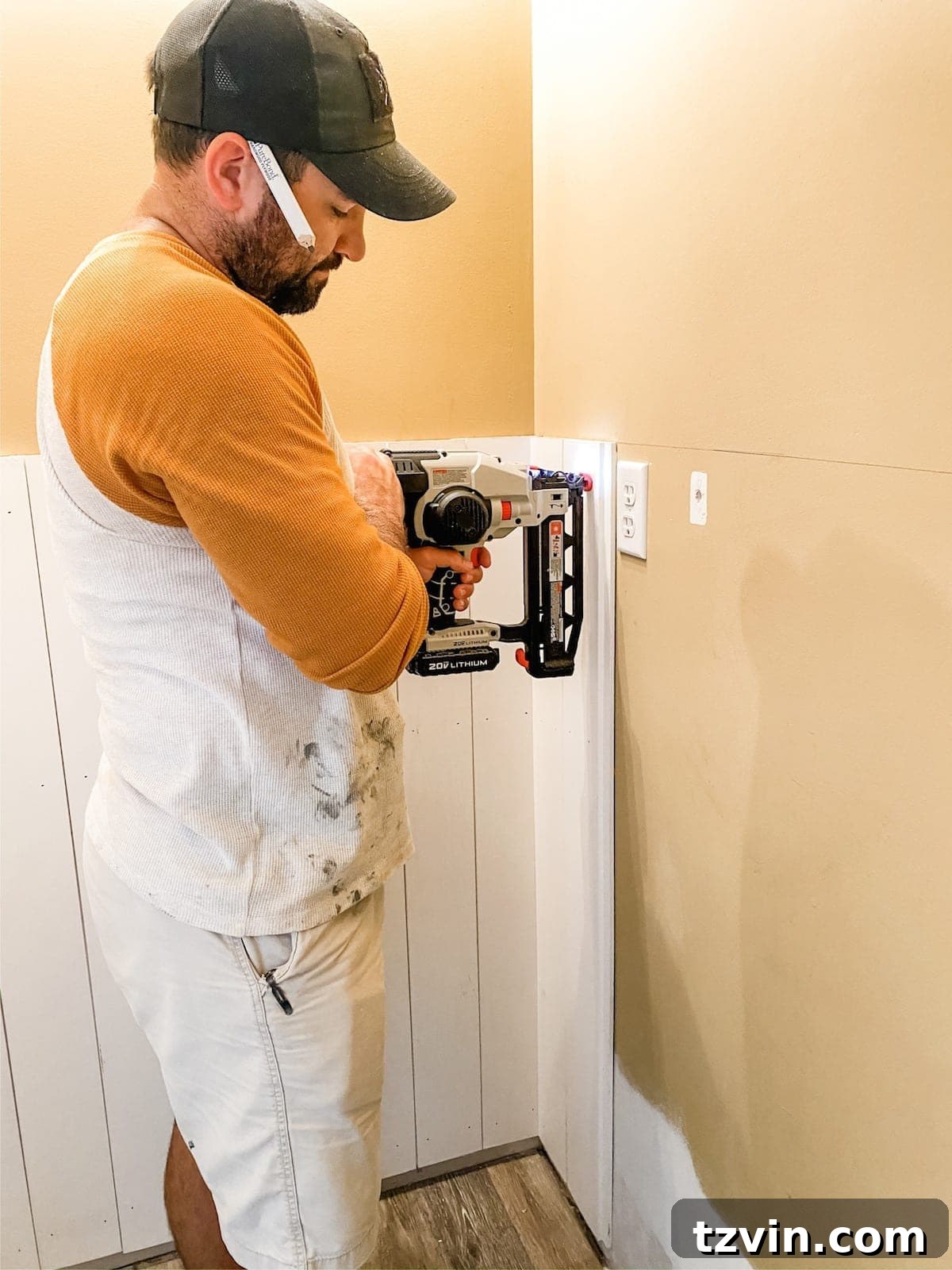 A close-up shot of a person using a cordless nailer to secure a vertical shiplap board to a bathroom wall, with the board already aligned and partially adhered.