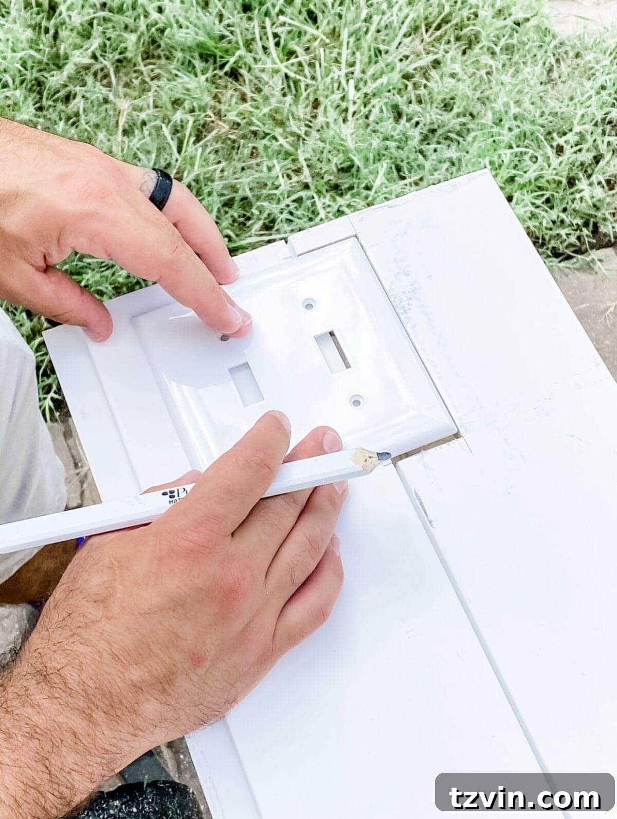 A hand tracing a standard outlet cover onto a shiplap board with a pencil, preparing the board for a precise cutout for electrical access.