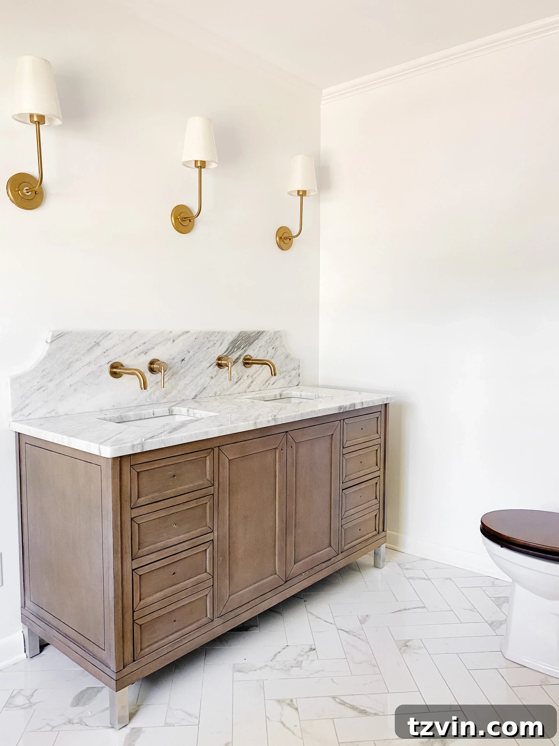 En Suite Bathroom Renovation: Faucets Are Finally Here! 2 Classic en suite bathroom featuring a scalloped marble backsplash, gold wall-mount sink faucets, faux marble porcelain tile herringbone floor, and a warm wooden vanity.
