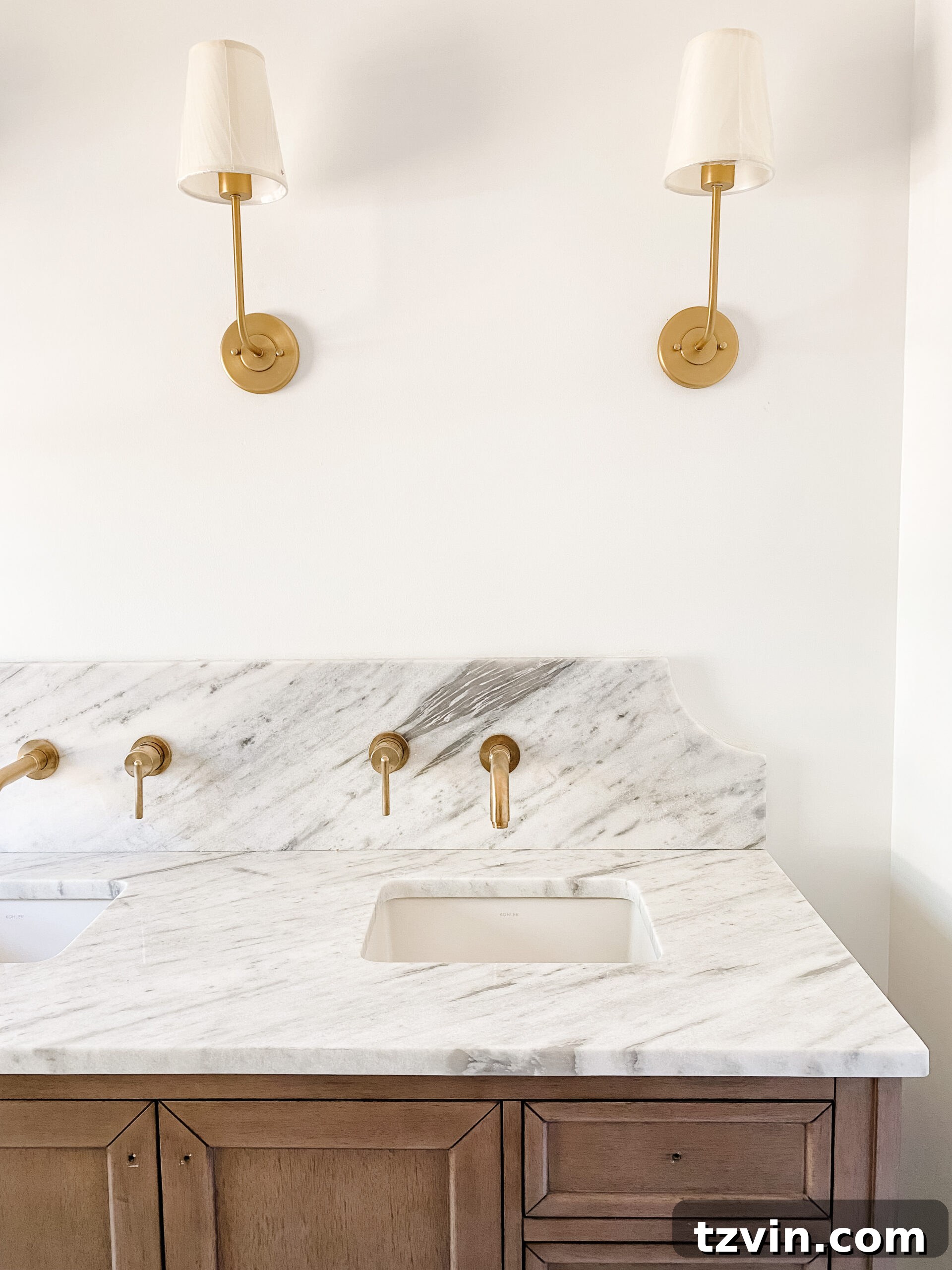 En Suite Bathroom Renovation: Faucets Are Finally Here! 3 Close-up of a scalloped marble backsplash with a gold wall-mount faucet and a classic wall sconce in an en suite bathroom.