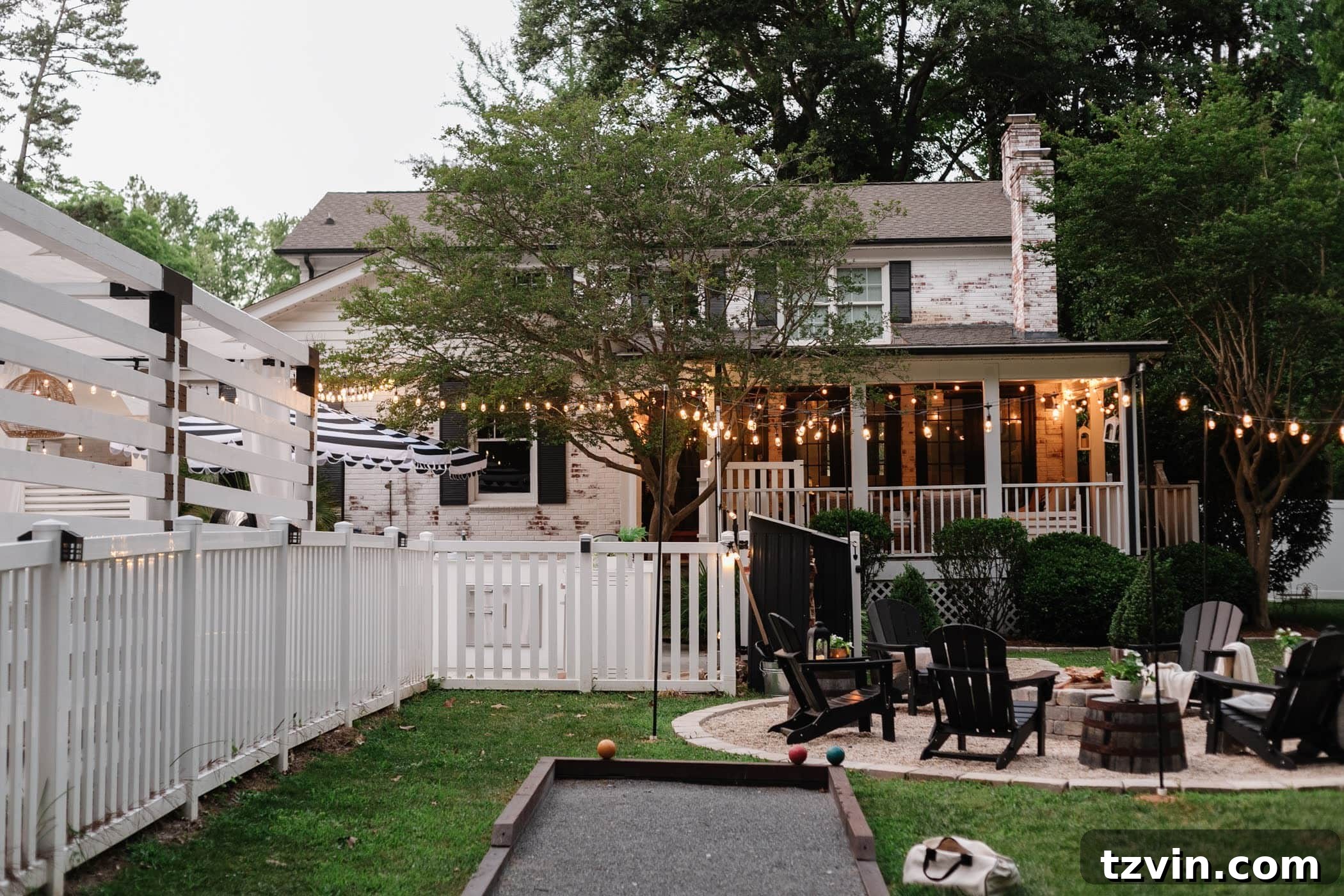 A beautifully transformed backyard featuring a limewashed brick house, a cozy fire pit area, a fun bocce ball court, a pristine white fence, a welcoming back porch, and charming string lights, creating an ideal space for outdoor entertainment.