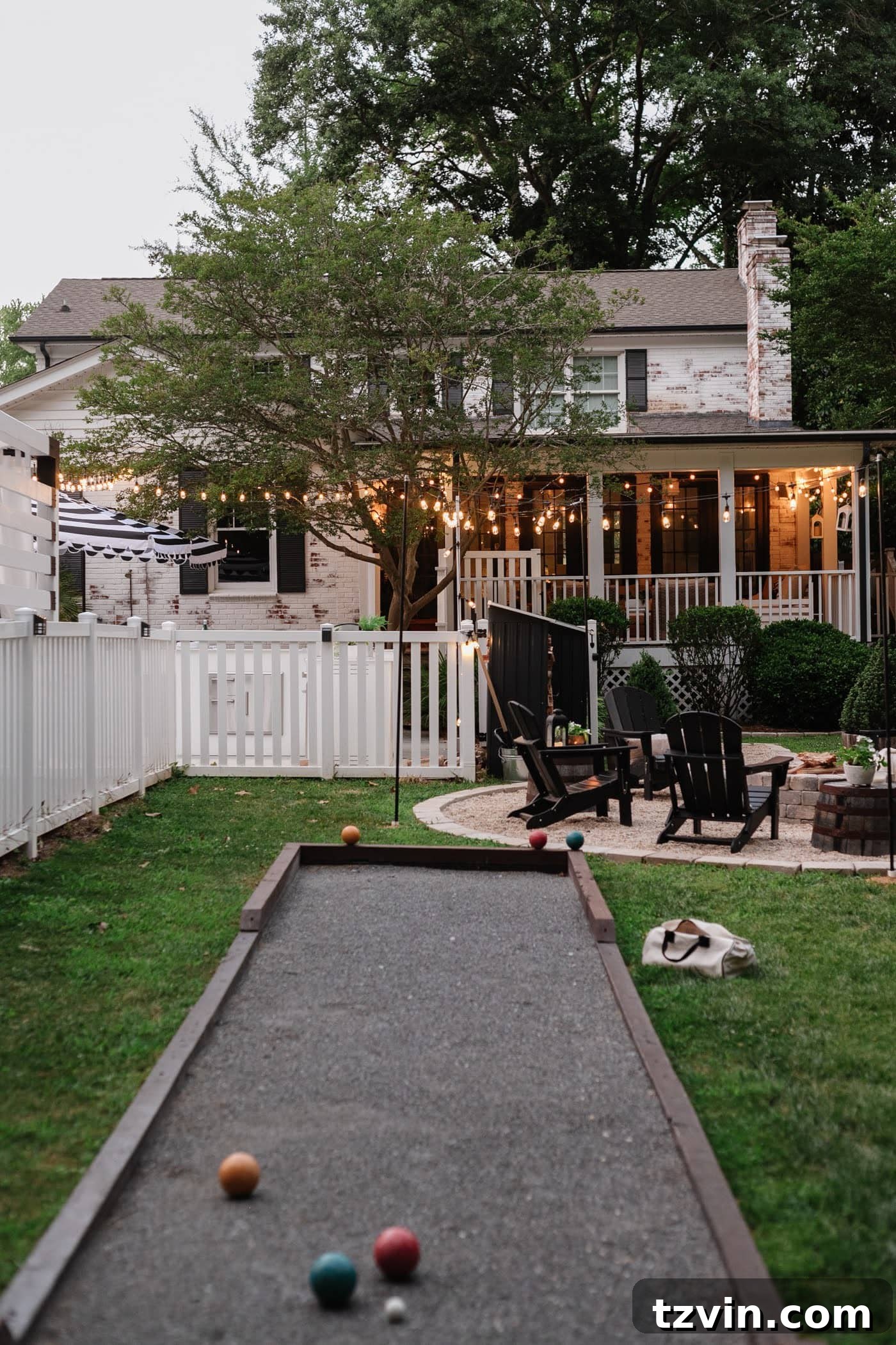 A beautifully transformed backyard featuring a limewashed brick house, a cozy fire pit area, a fun bocce ball court, a pristine white fence, a welcoming back porch, and charming string lights, creating an ideal space for outdoor entertainment.