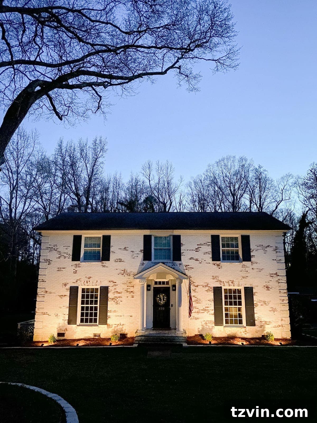 A beautifully limewashed brick colonial house illuminated by warm solar spotlights, showcasing its enhanced curb appeal and inviting ambiance at dusk.