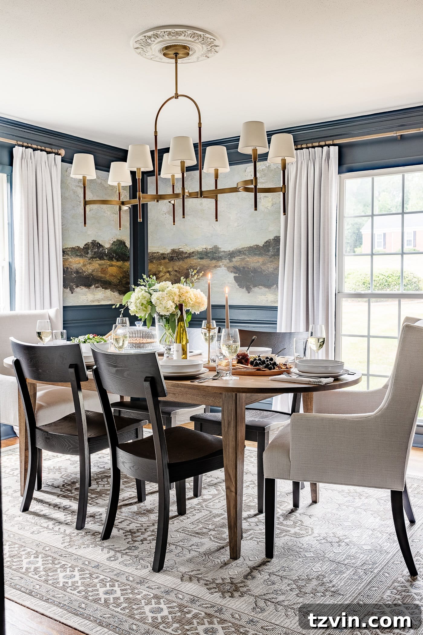 A stunning navy blue dining room featuring captivating mural wallpaper framed by elegant picture frame molding, luxurious linen curtains, a chic shaded chandelier, a beautiful wood oval dining table, sophisticated linen head chairs, and stylish black dining chairs, creating a dramatic and inviting atmosphere.