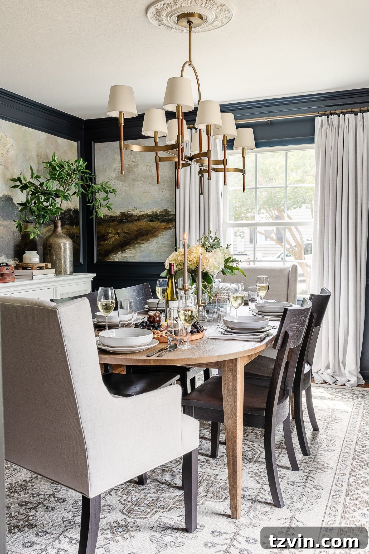 A stunning navy blue dining room featuring captivating mural wallpaper framed by elegant picture frame molding, luxurious linen curtains, a chic shaded chandelier, a beautiful wood oval dining table, sophisticated linen head chairs, and stylish black dining chairs, creating a dramatic and inviting atmosphere.
