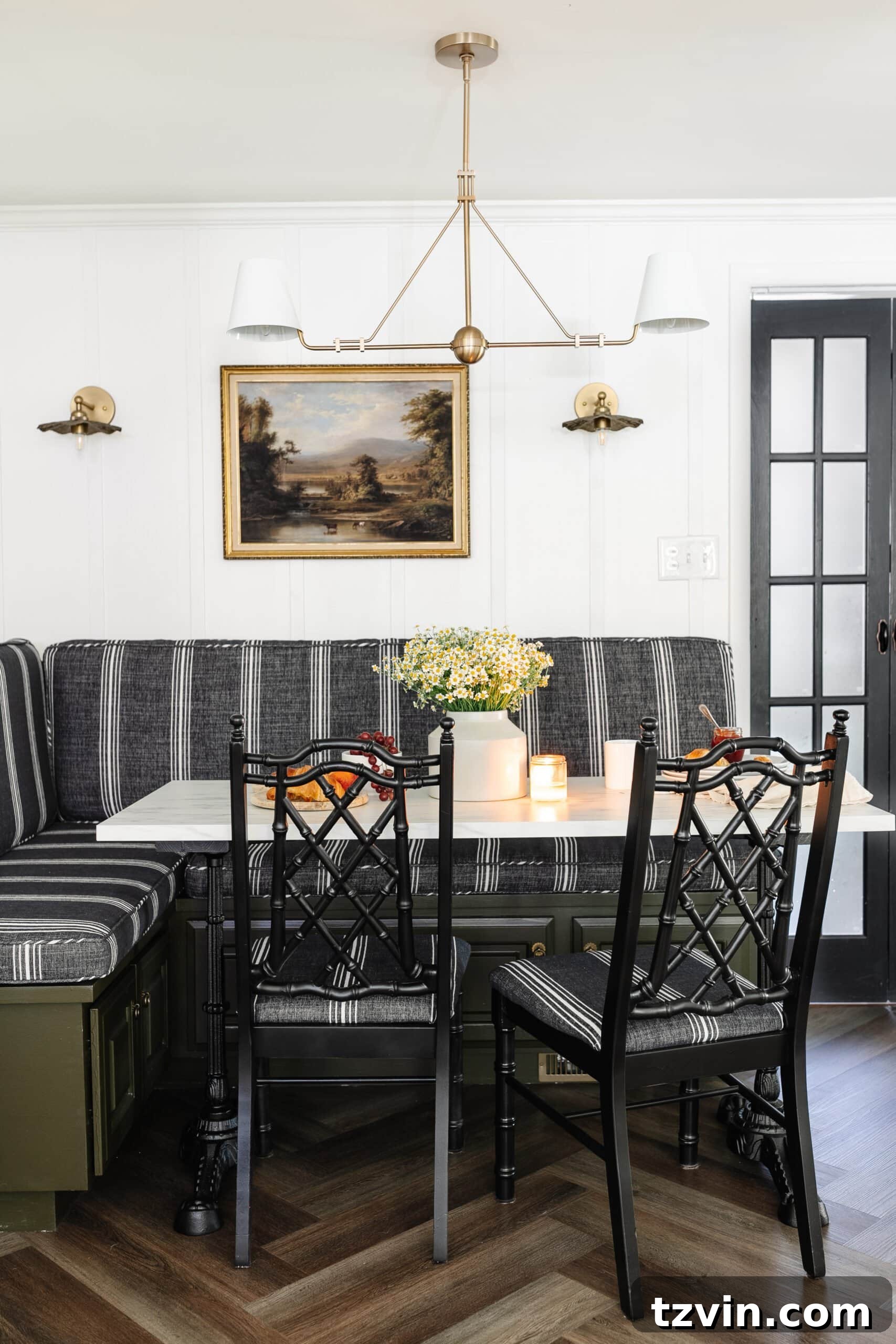 A charming cottagecore breakfast nook, featuring a built-in banquette, an elegant shaded chandelier, a practical bifold French door, stylish herringbone floor, and a sophisticated marble bistro table, creating a cozy and functional dining area.