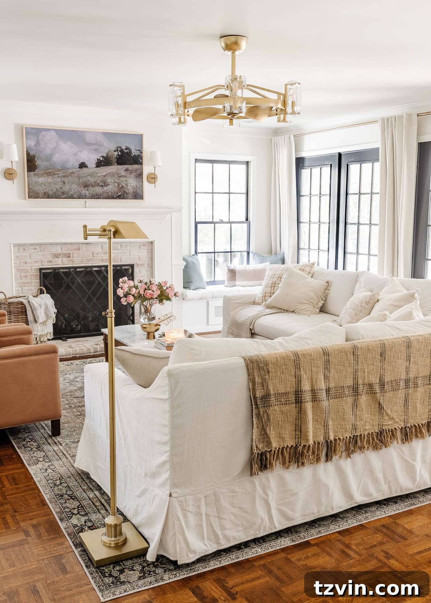 A bright white living room featuring a cozy limewashed brick fireplace, an elegant gold fandelier, comfortable white Pottery Barn sofas, luxurious leather recliners, a discreet art TV, and sophisticated black French doors, creating a serene and inviting atmosphere.