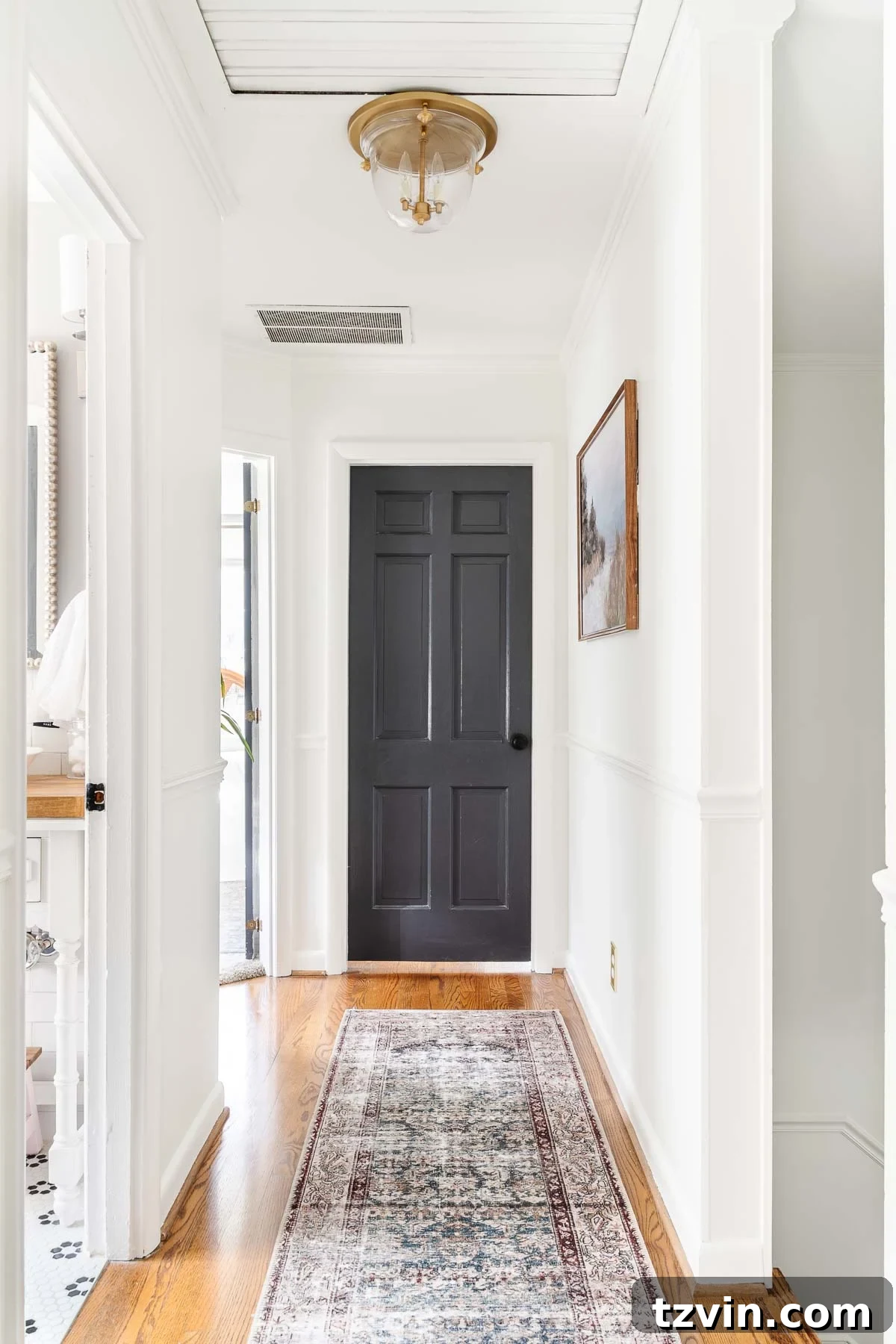 A bright white hallway featuring contrasting black doors, a vintage runner rug, an elegant brass light fixture, and charming landscape art, creating a stylish and inviting transitional space.