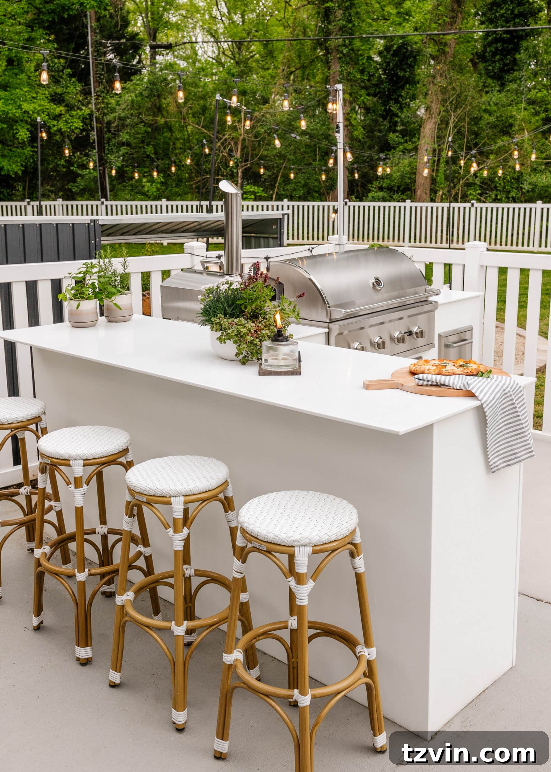 Spring Patio Refresh 8 white outdoor kitchen with grill, bar stools, string lights, white fence, and potted plants