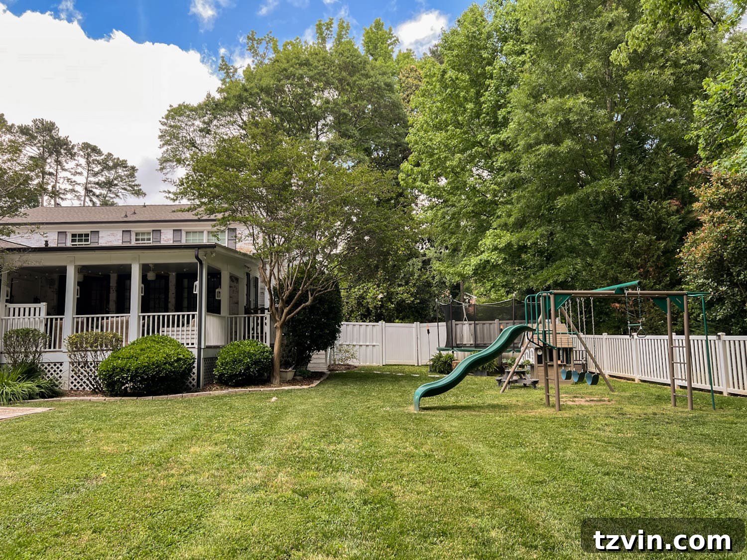 Stepping stones lead to an inviting outdoor trampoline, flanked by lush ferns and atmospheric tiki torches, near a charming playhouse, picnic table, and swing set in a revitalized side yard play area.