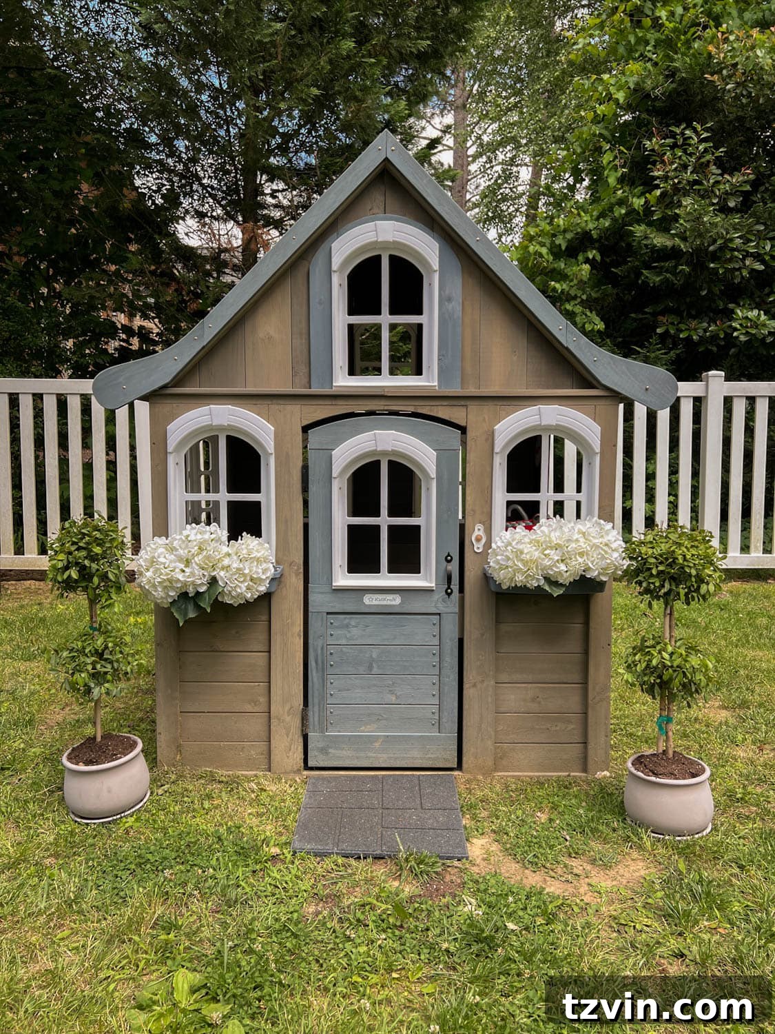 Charming wooden outdoor playhouse adorned with white faux hydrangeas in window boxes and topiaries, creating a whimsical focal point in the side yard play area.
