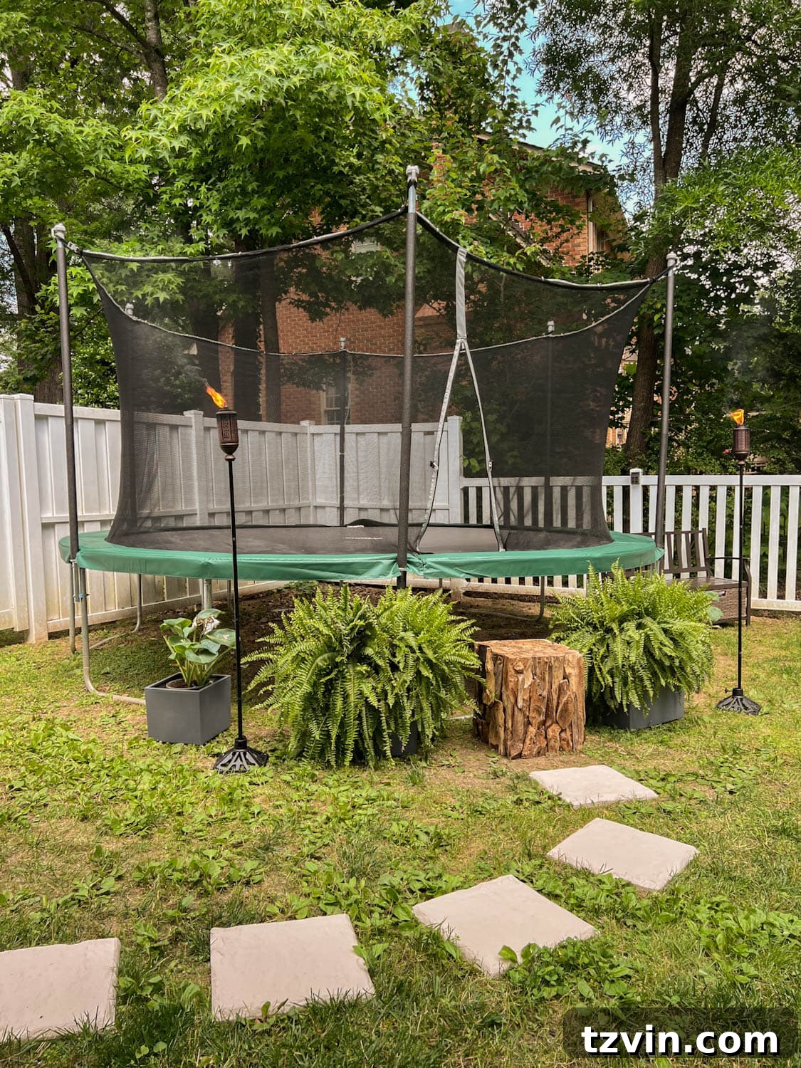 Beautifully laid stepping stones leading to an outdoor trampoline, complemented by a natural tree stump step, lush ferns, and atmospheric tiki torches in a lively side yard play area.