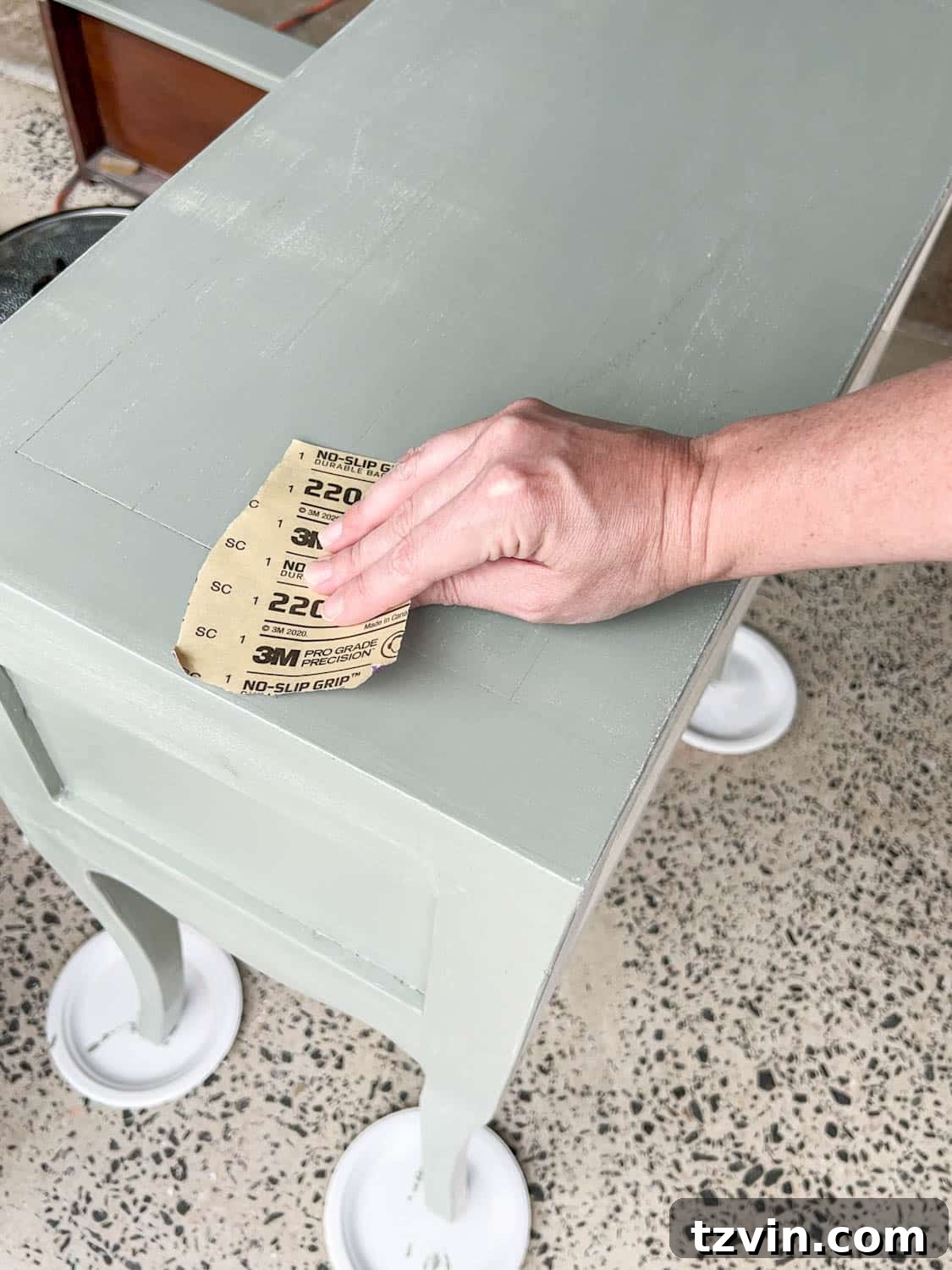 A hand lightly sanding a painted nightstand with ultra-fine sandpaper between coats to achieve a perfectly smooth and flawless gloss finish.