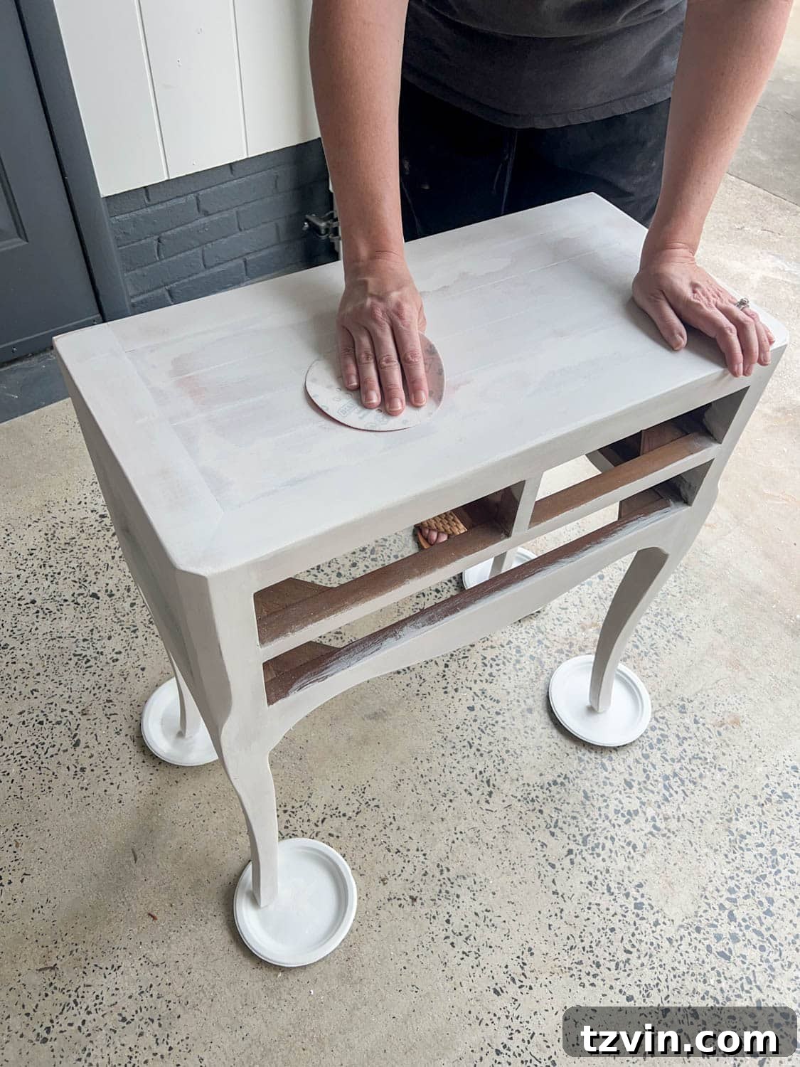 A hand gently sanding a primed furniture surface with ultra-fine sandpaper, a key step for achieving a perfectly smooth foundation before applying gloss paint.