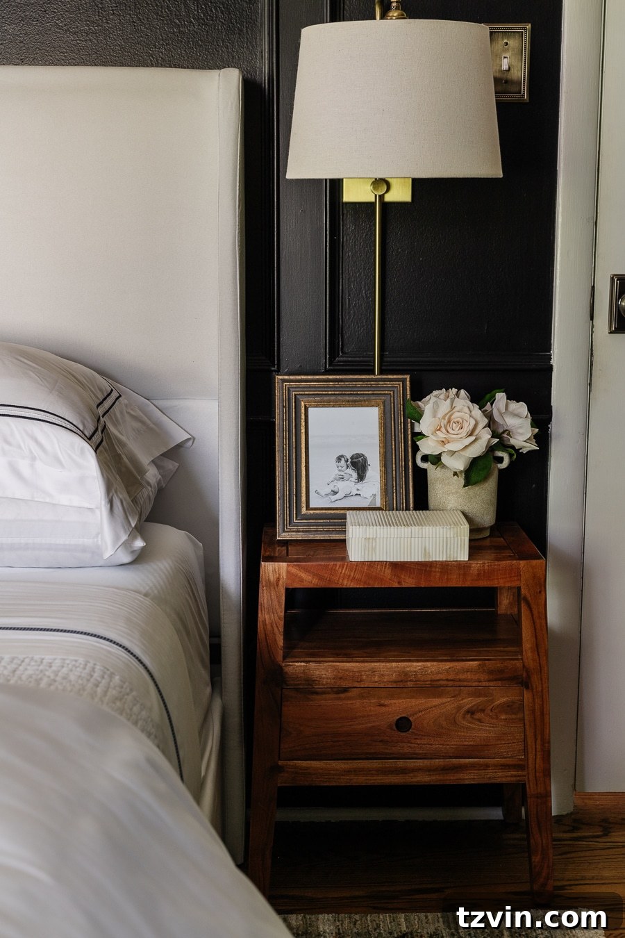 wooden nightstand beside bed with sconce lamp flowers and picture frame